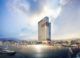 Un moderno hotel de gran altura con vista al agua, que cuenta con varios pisos con balcones, un puerto deportivo con barcos y un paisaje urbano de fondo.