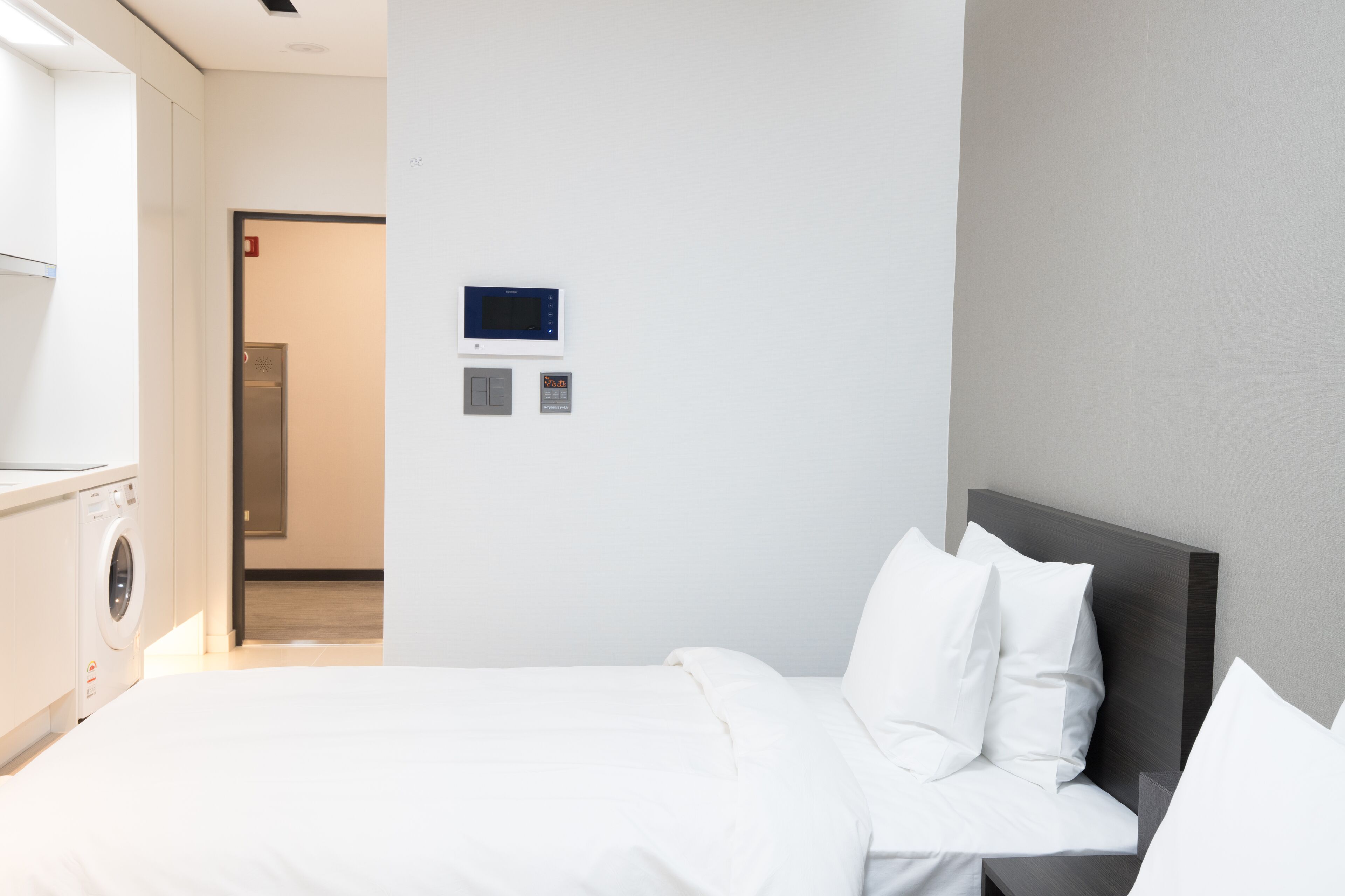A hotel room with a bed, a washing machine, and a control panel on the wall.