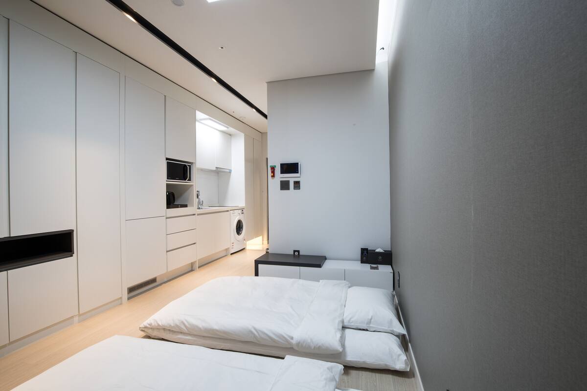 A modern hotel room with two beds, a kitchenette, and a small bathroom.