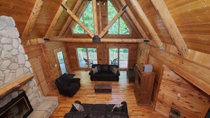A log cabin interior with a stone fireplace, two black sofas, a wooden coffee table, and a view of the deck through large windows. - Spacious 8-Bedroom Retreat at Martinwoods Cabins, Perfect for family gatherings (Rockbridge)