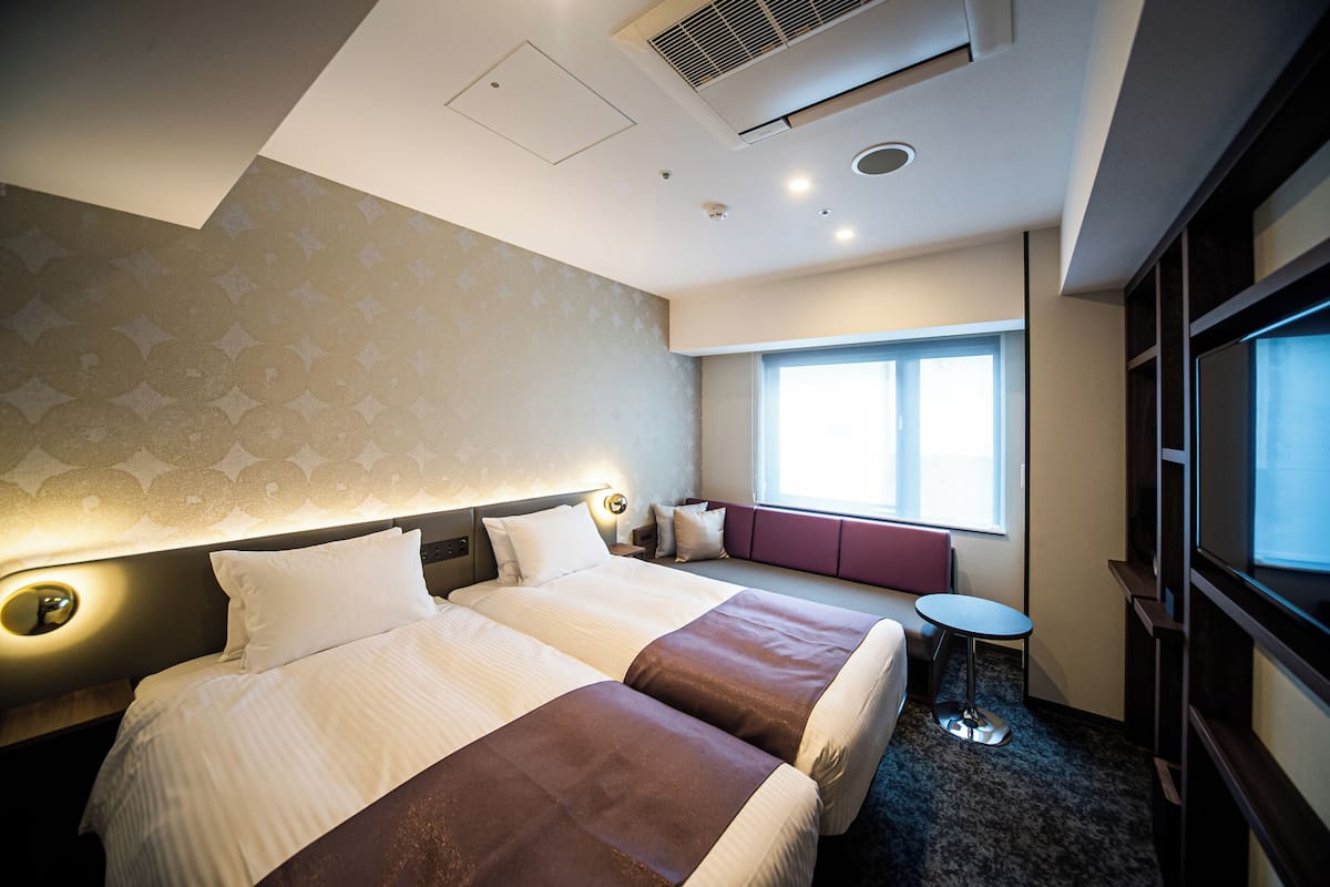 A hotel room with two beds, a small table, a seating area by the window, and a TV.