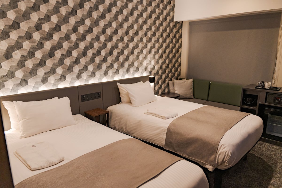 A hotel room with two beds, a headboard with a geometric pattern, a bedside table with a lamp, and a small seating area with a green cushion.