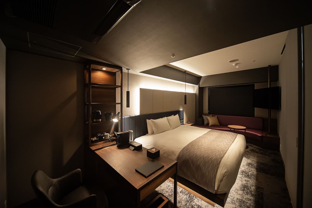 A modern hotel room with a large bed, a desk with a chair, a small table, and a sofa.