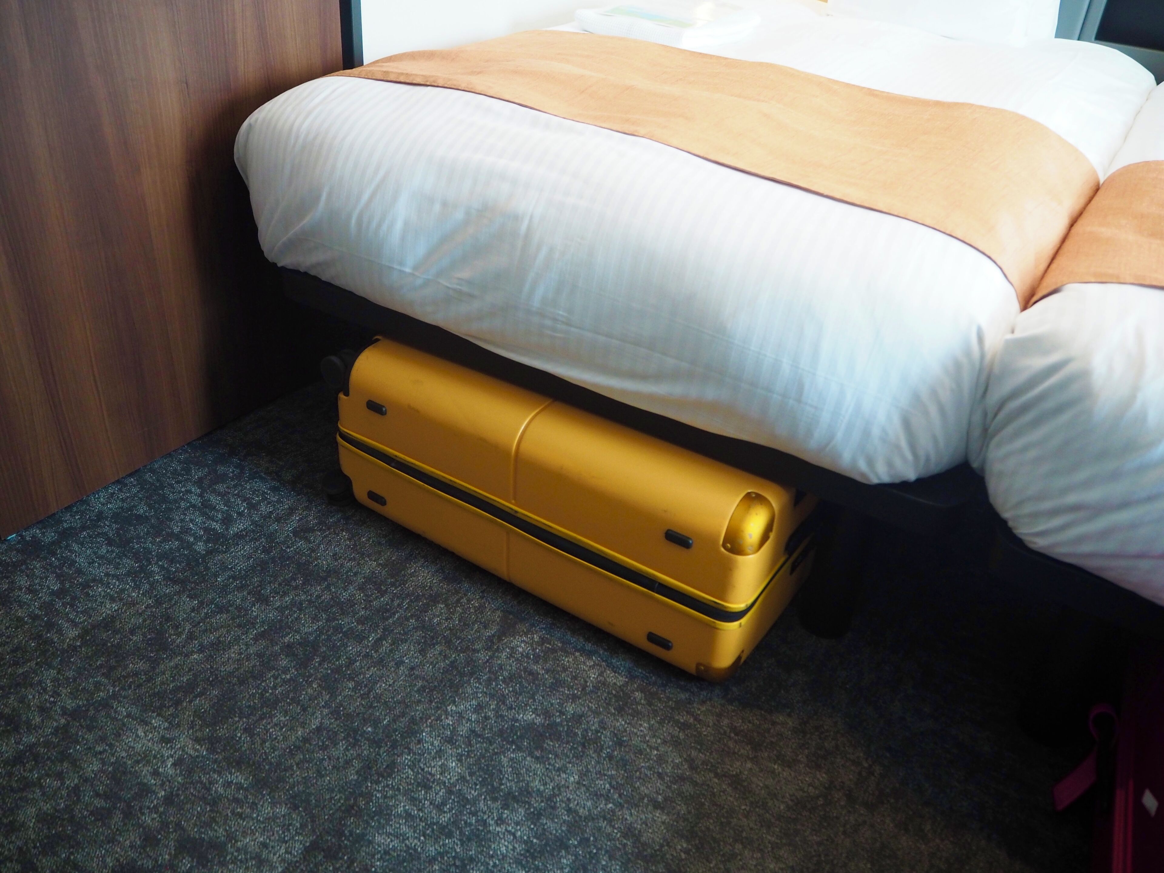 A bed with a white and orange bedspread, a yellow suitcase under the bed, and a dark carpeted floor.