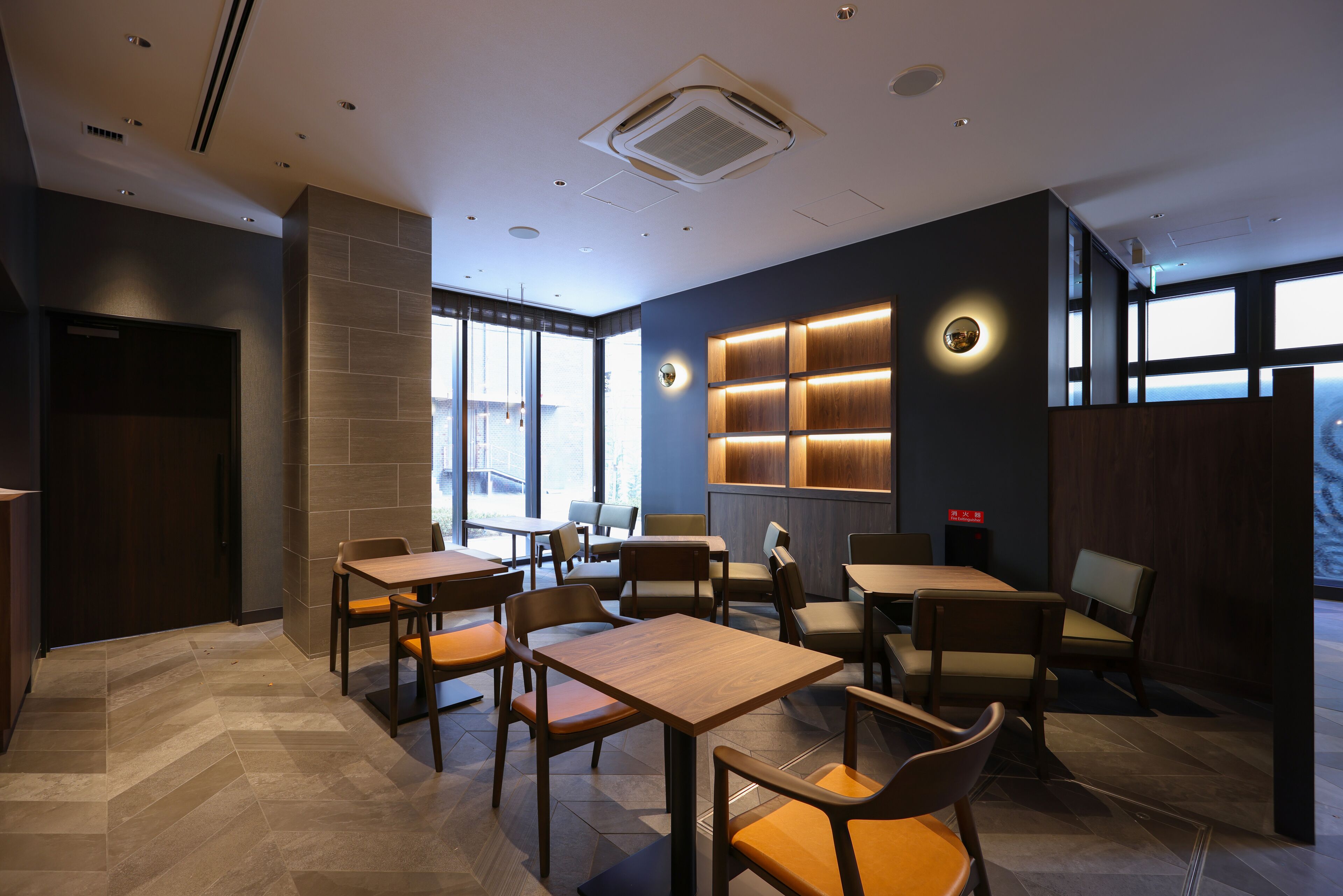 Un interior de restaurante moderno con varias mesas y sillas, un ventanal grande y una lámpara fijada a la pared.