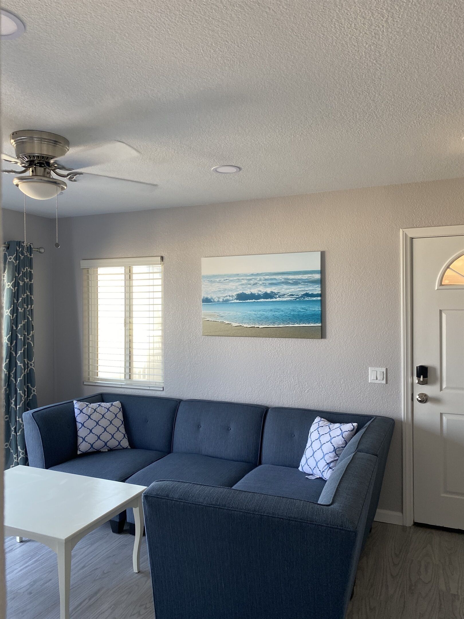 Un salon avec un canapé bleu foncé, une table basse blanche, un ventilateur de plafond, une fenêtre avec des stores et un tableau représentant l’océan.
