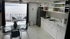 Une cuisine moderne avec vue sur la mer, un coin repas doté d’une table en verre et des meubles de rangement blancs.
