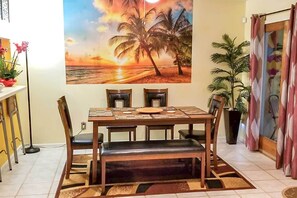 A dining area with a wooden table, four chairs, and a bench. There is a painting of a sunset over palm trees on the wall behind the table.