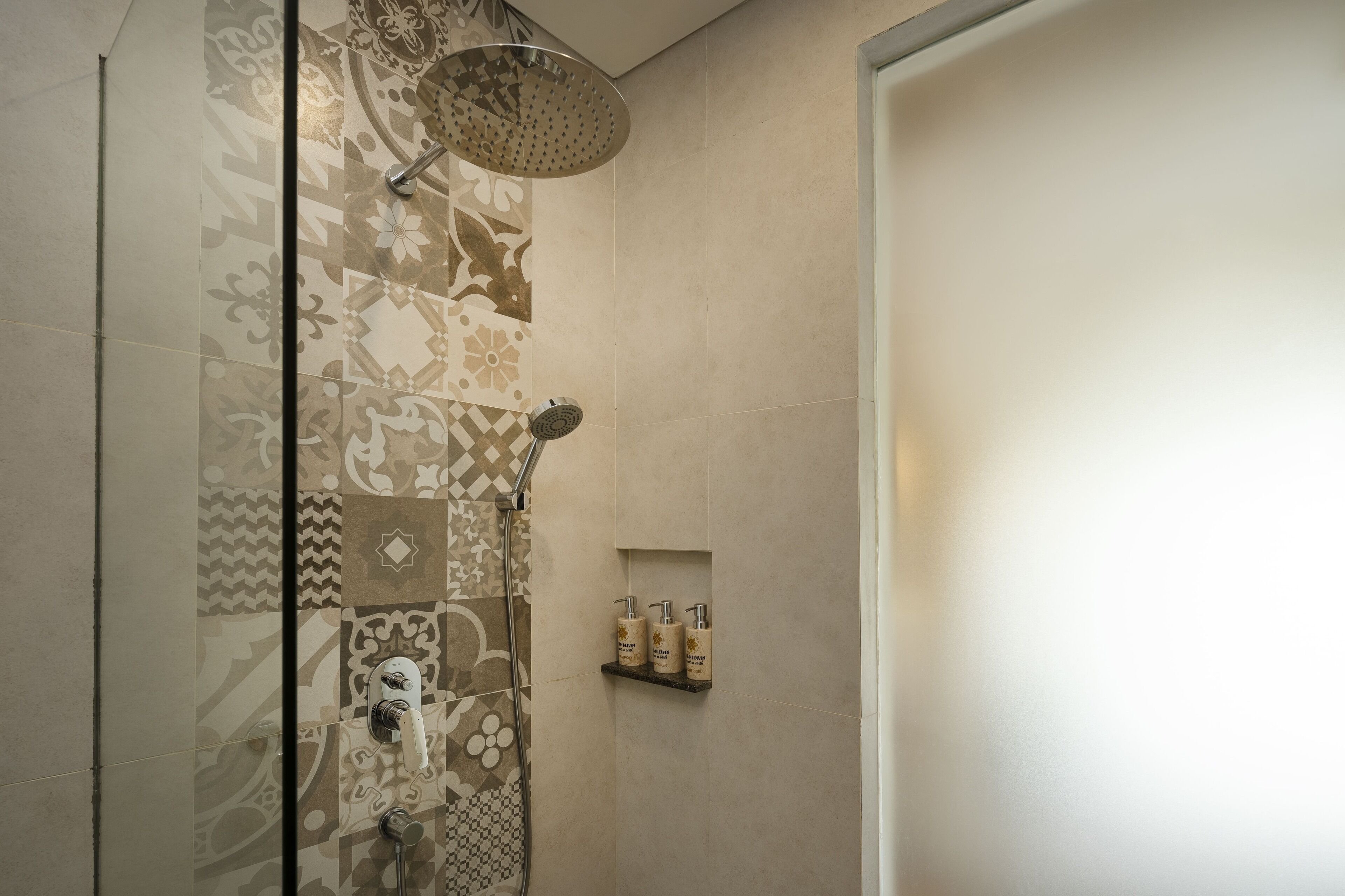 A shower with a rain showerhead and a handheld showerhead, a wall-mounted shelf with toiletries, and a door to the right.