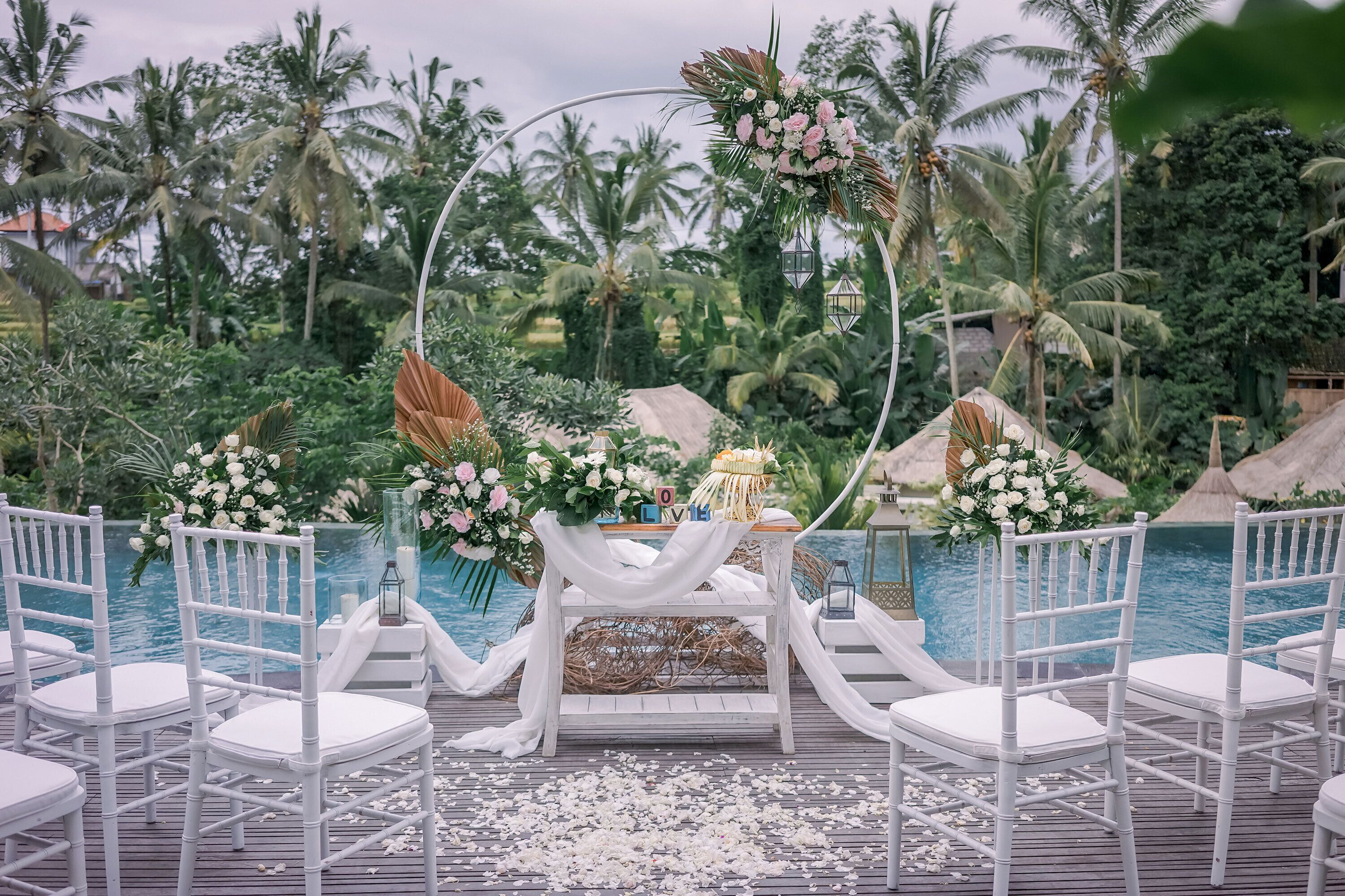 Une cérémonie de mariage est mise en place avec des chaises blanches et un autel en bois, décoré de fleurs et de lanternes, donnant sur une piscine bordée de palmiers.