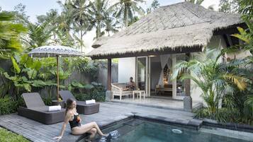 Un área junto a la piscina con sillones de descanso, una estructura de techo de paja y exuberante vegetación tropical.