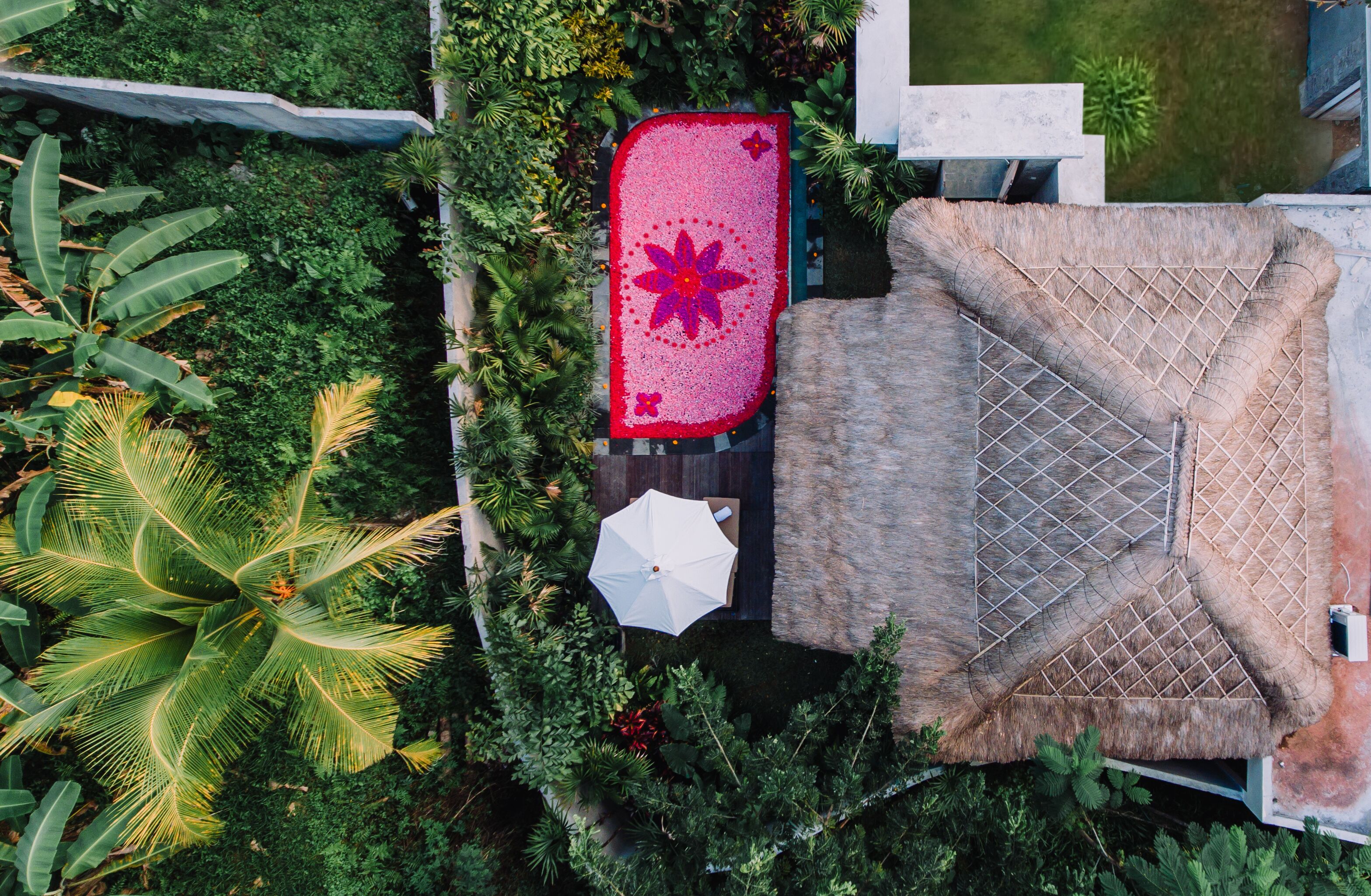 An aerial view of a resort with a thatched roof structure, a red mat with a flower design, and lush tropical vegetation.