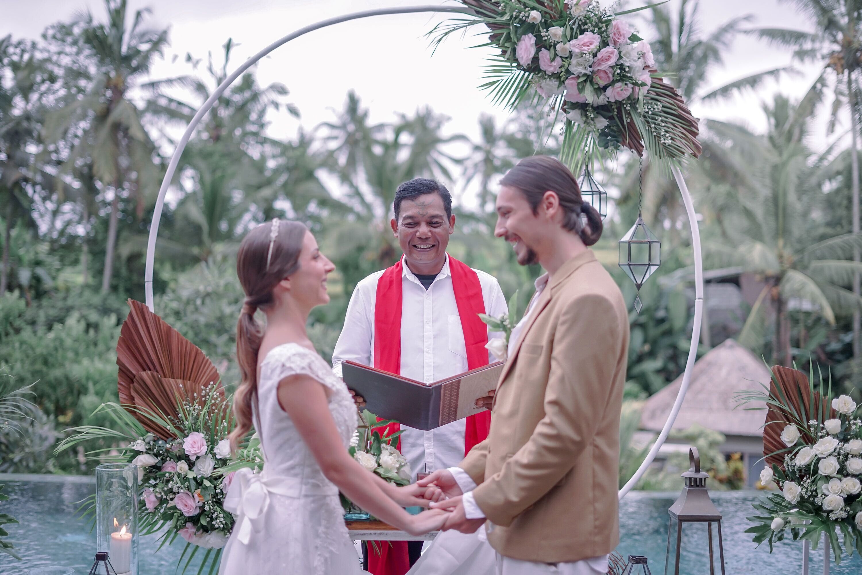 A couple exchanging rings under a floral arch with a pool and palm trees in the background.