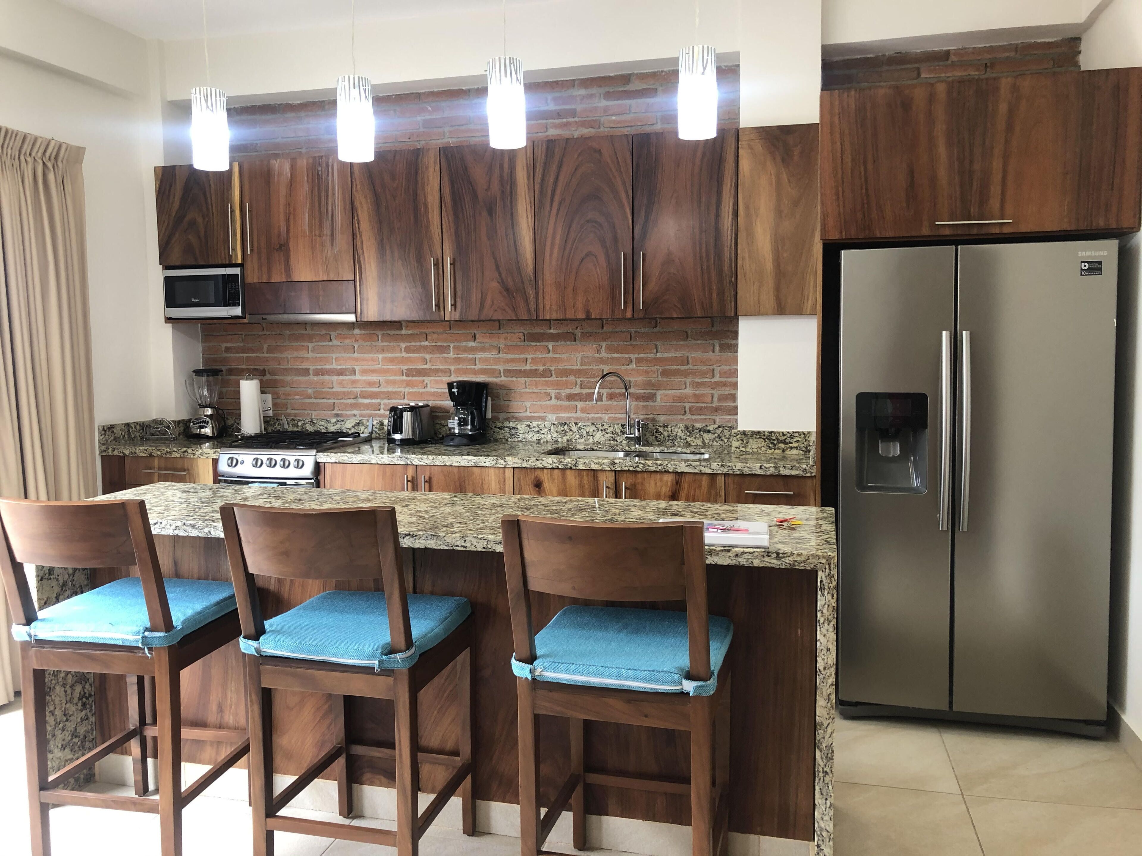 A modern kitchen with wooden cabinets, granite countertops, a stainless steel refrigerator, and bar stools with blue cushions.