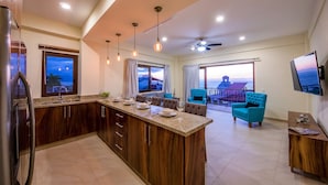 A modern kitchen with a central island, dining area with blue chairs, a TV mounted on the wall, and a balcony with a view. - Nise (Puerto Vallarta)