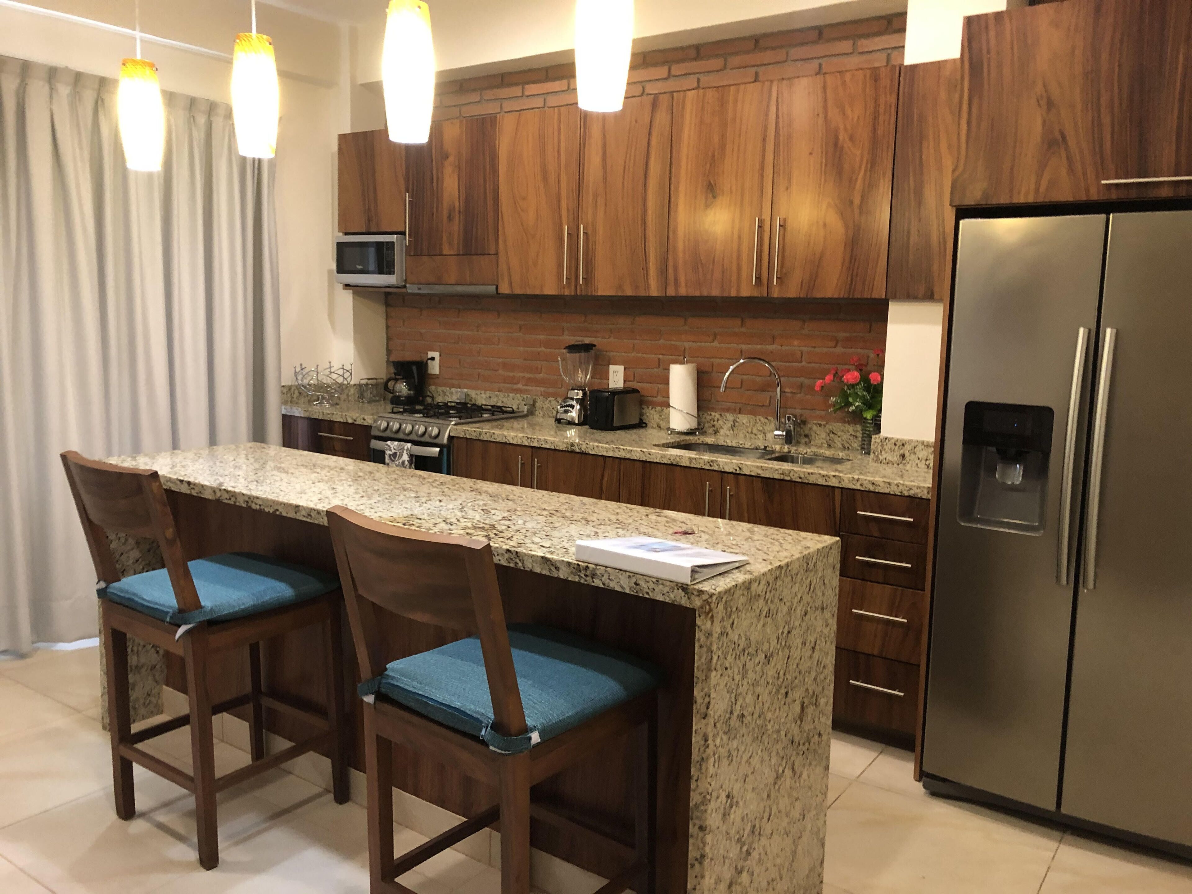 A modern kitchen with granite countertops, wooden cabinets, a stainless steel refrigerator, and two bar stools with blue cushions.