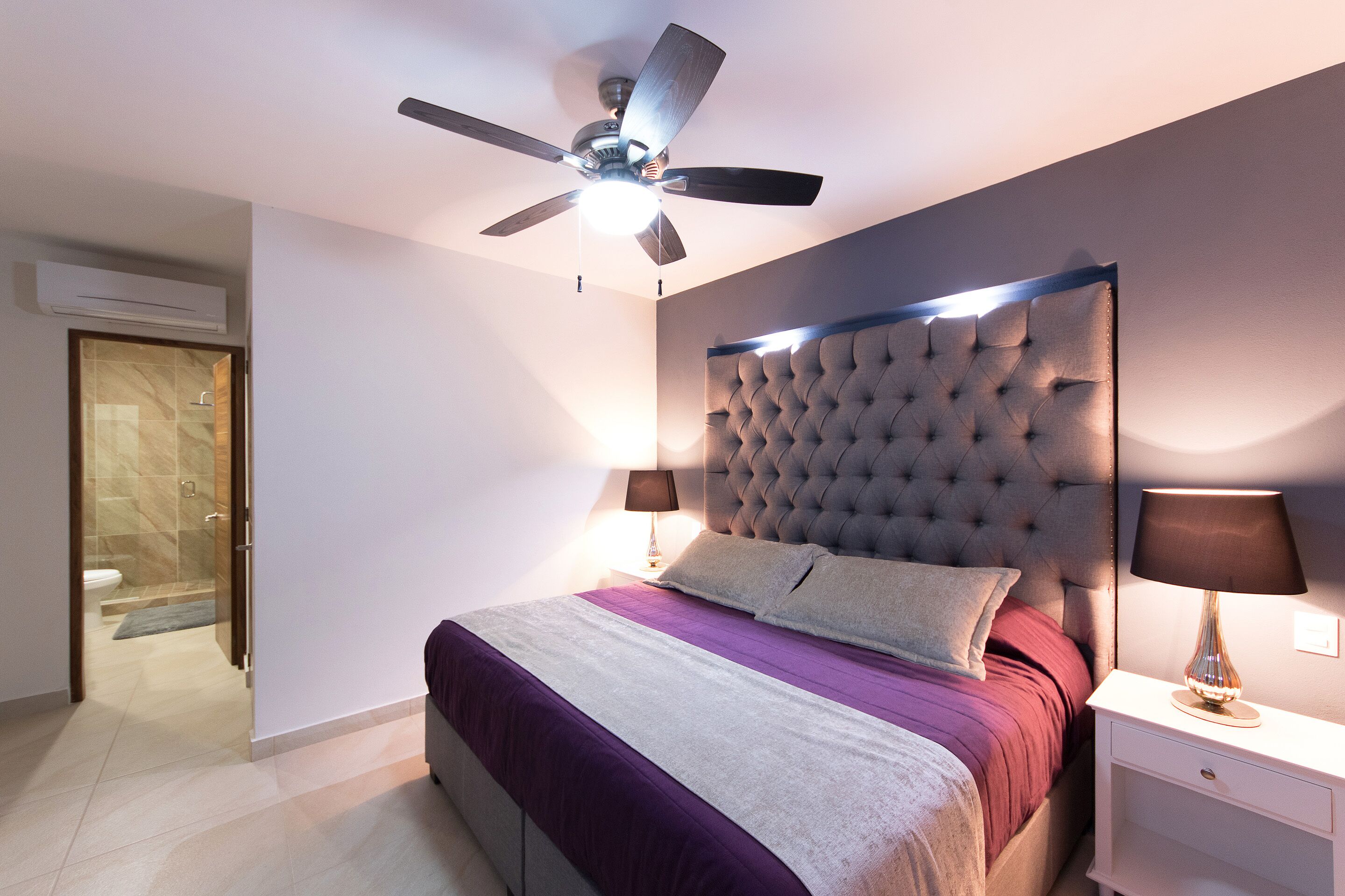 Une chambre moderne avec une tête de lit capitonnée, un ventilateur de plafond, une lampe de chevet et une vue sur la salle de bain.