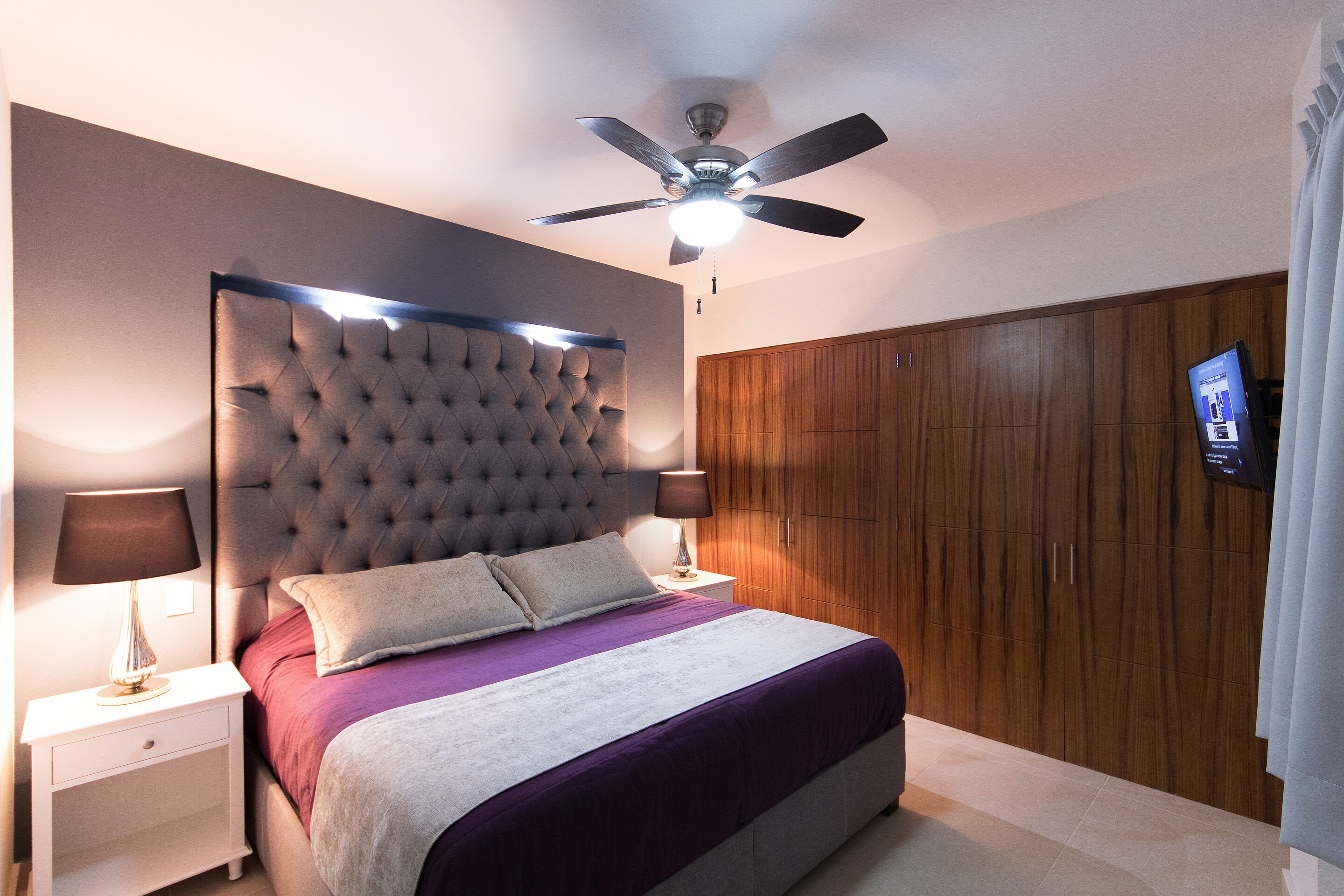 A bedroom with a tufted headboard, a bed with a purple and white bedspread, two bedside lamps, a ceiling fan, wooden wardrobe, and a television on the wall.