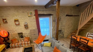 A cozy living room with a grey sofa, wooden coffee table, and orange chairs. A stone wall and wooden beams are visible in the background.