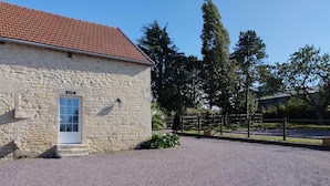 A stone building with a red-tiled roof, a gravel driveway, and a fenced area with trees in the background.