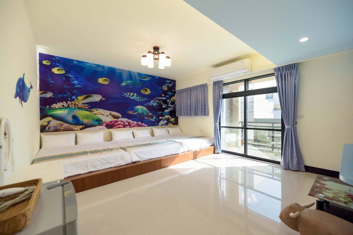 A hotel room with a large mural of an underwater scene, a bed with white linens, a large window with curtains, and a small table with a chair.