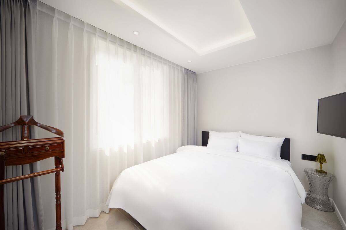 A modern hotel room with a large bed, a wooden dresser, a bedside table with a lamp, and a large window with sheer curtains.