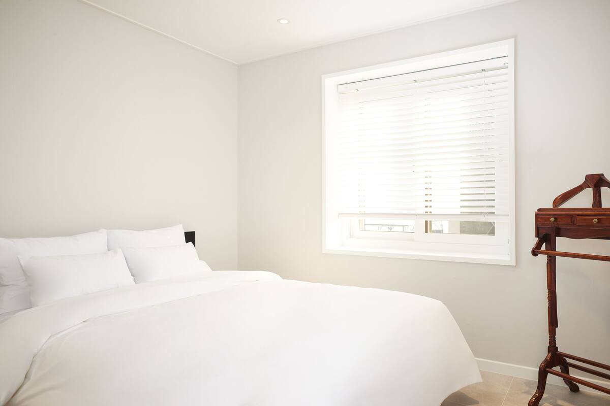 A bedroom with a bed, a window with blinds, and a wooden nightstand.