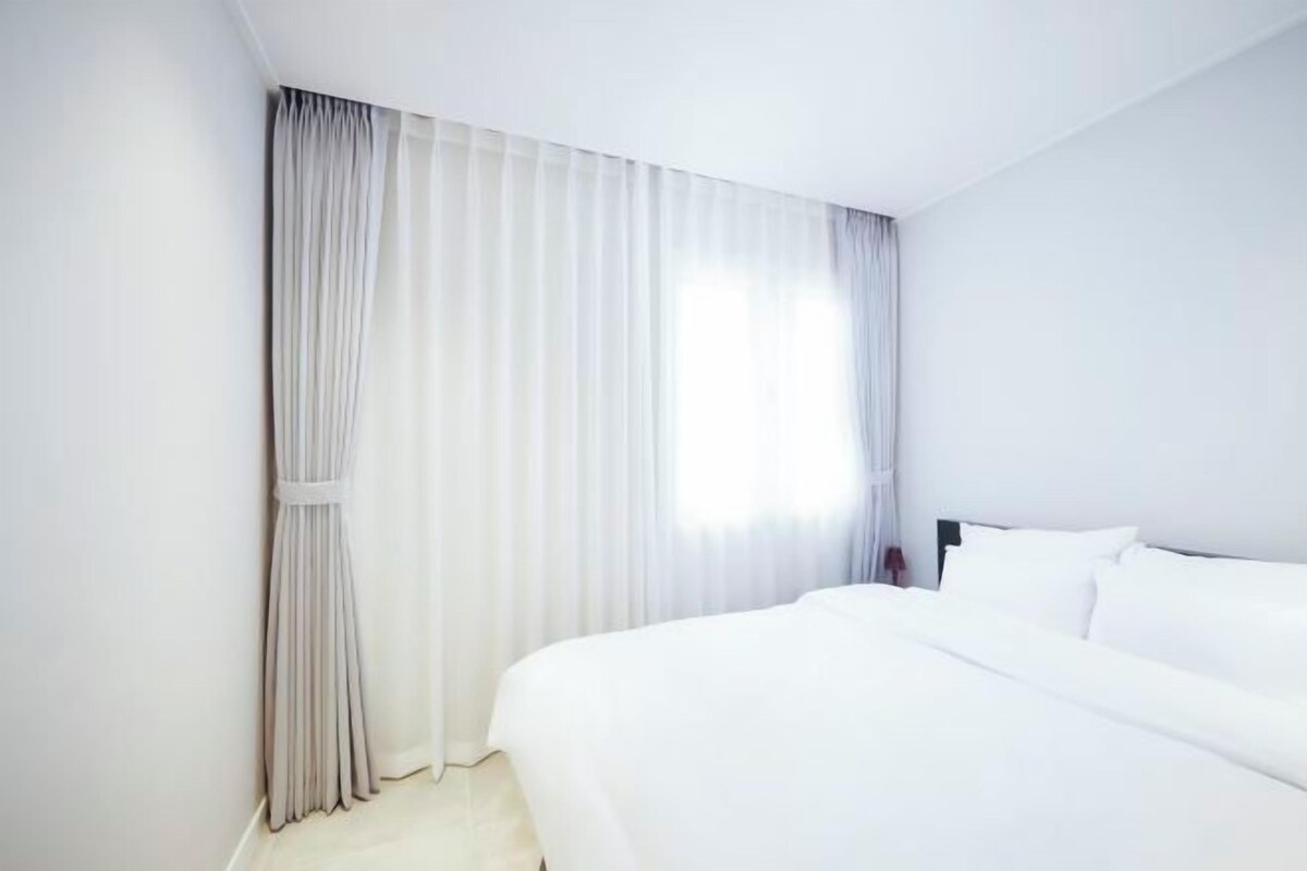 Quarto de hotel com cama grande, cortinas brancas e janela com luz natural.