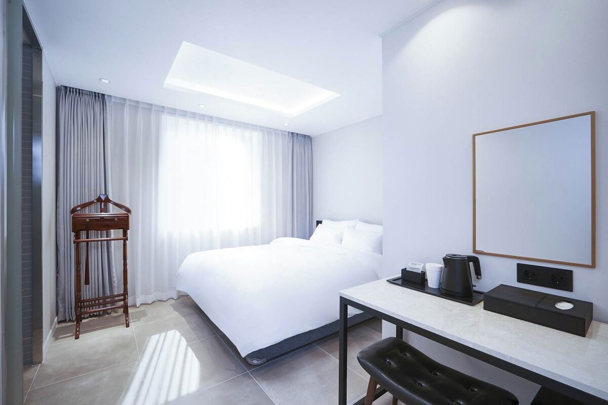 A hotel room with a bed, a desk with a chair, a hanging coat rack, and a large mirror.