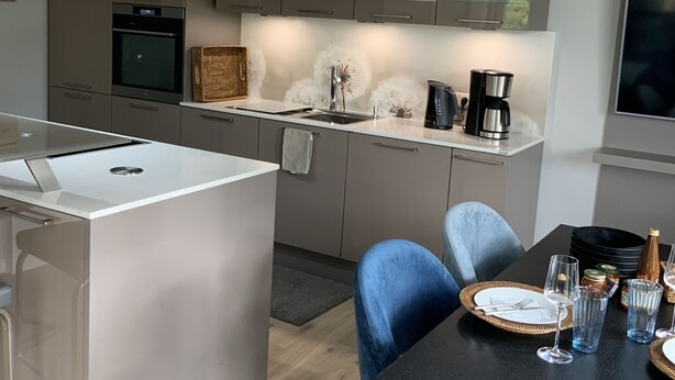 A modern kitchen with a central island, built-in oven, and a dining area with a black table and blue chairs. - Exclusive apartment with private sauna, high-quality kitchen (Zingst)
