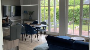 A modern living room with a blue sofa, a dining area with a table and chairs, a TV mounted on the wall, and large windows offering a view of greenery outside. - Exclusive apartment with private sauna, high-quality kitchen (Zingst)
