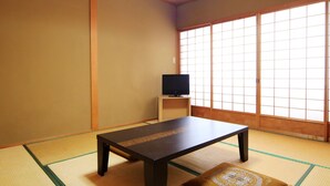 Une pièce de style japonais traditionnel, avec un sol recouvert de tatamis, une table basse, un téléviseur posé sur un meuble et des portes coulissantes.
