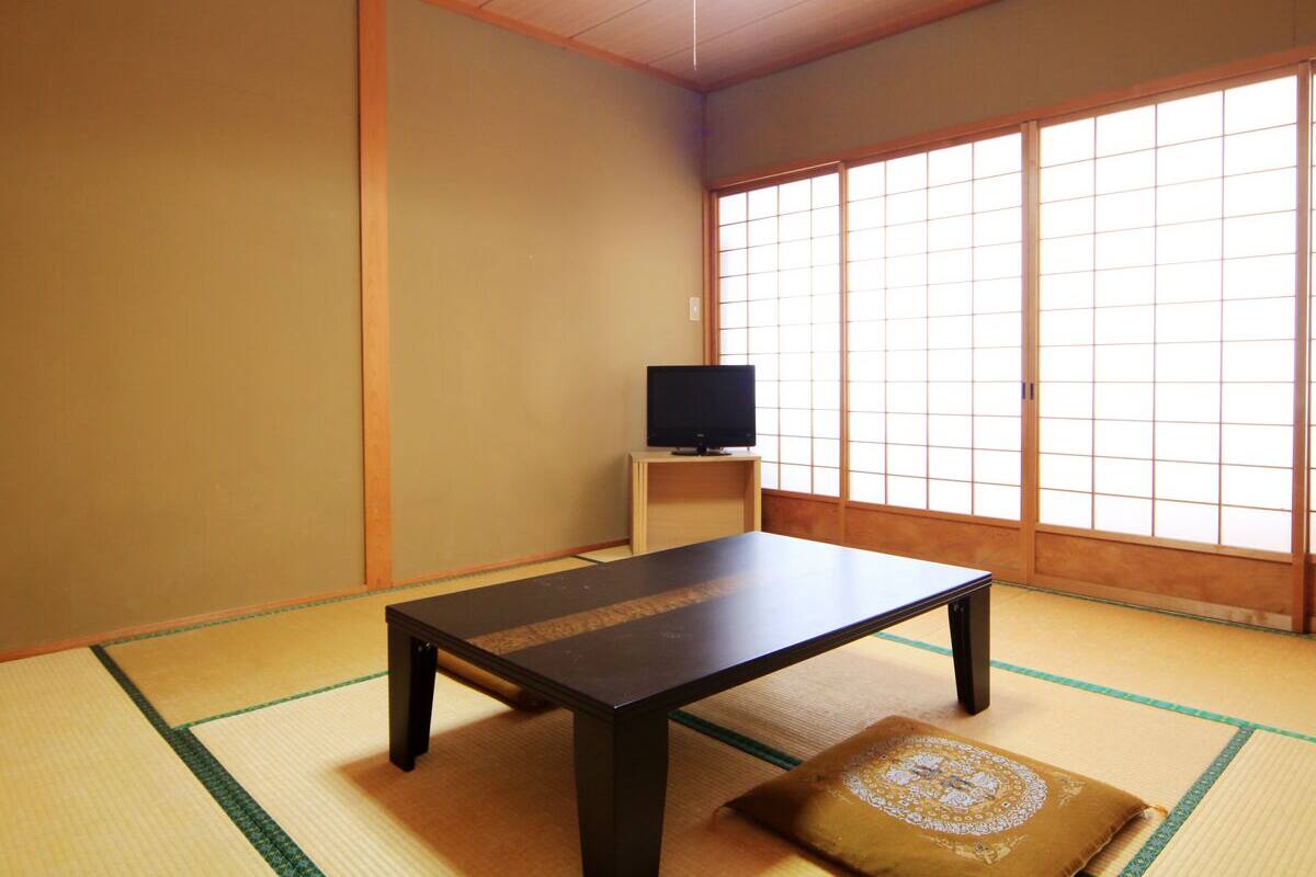 A traditional Japanese room with tatami flooring, a low table, a television on a stand, and sliding doors.