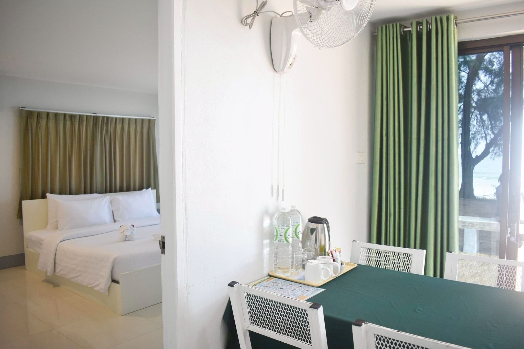 Une chambre d’hôtel avec un lit, une table à manger avec des chaises, un ventilateur et une fenêtre avec des rideaux.