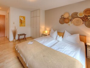 A neatly arranged bedroom with a large bed, a bedside table with a lamp, a wardrobe, and a wall decorated with woven baskets.