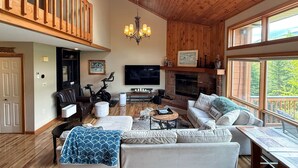 Un salón amplio con techo alto, vigas de madera, chimenea, televisor y un ventanal grande.