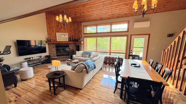 Amplia sala de estar con chimenea, televisor, comedor con una mesa larga y ventanas grandes con vistas al exterior.
