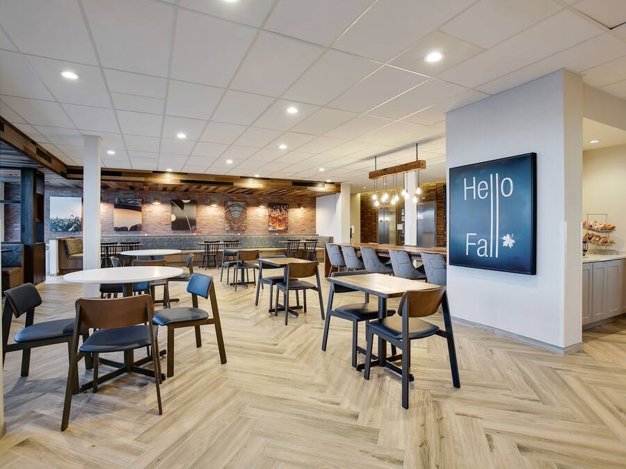 A modern dining area with multiple tables and chairs, a kitchen in the background, and a 'Hello Fall' sign on the wall.