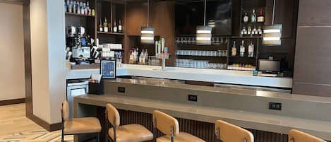 Uma área de bar moderna com um balcão comprido, vários bancos e uma variedade de garrafas e copos nas prateleiras.
