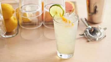 Ein Glas helles Getränk mit Limettenscheibe und Orangenscheibe, umgeben von leeren Gläsern und Zitronen.