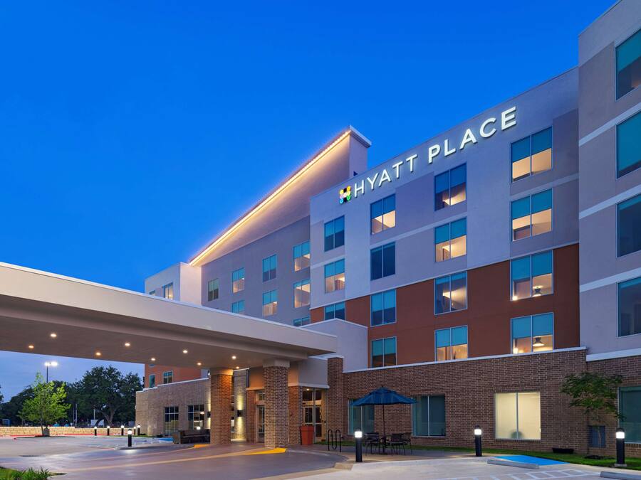Un hotel de varios pisos con un llamativo letrero de "HYATT PLACE", una entrada cubierta y una zona de piscina.