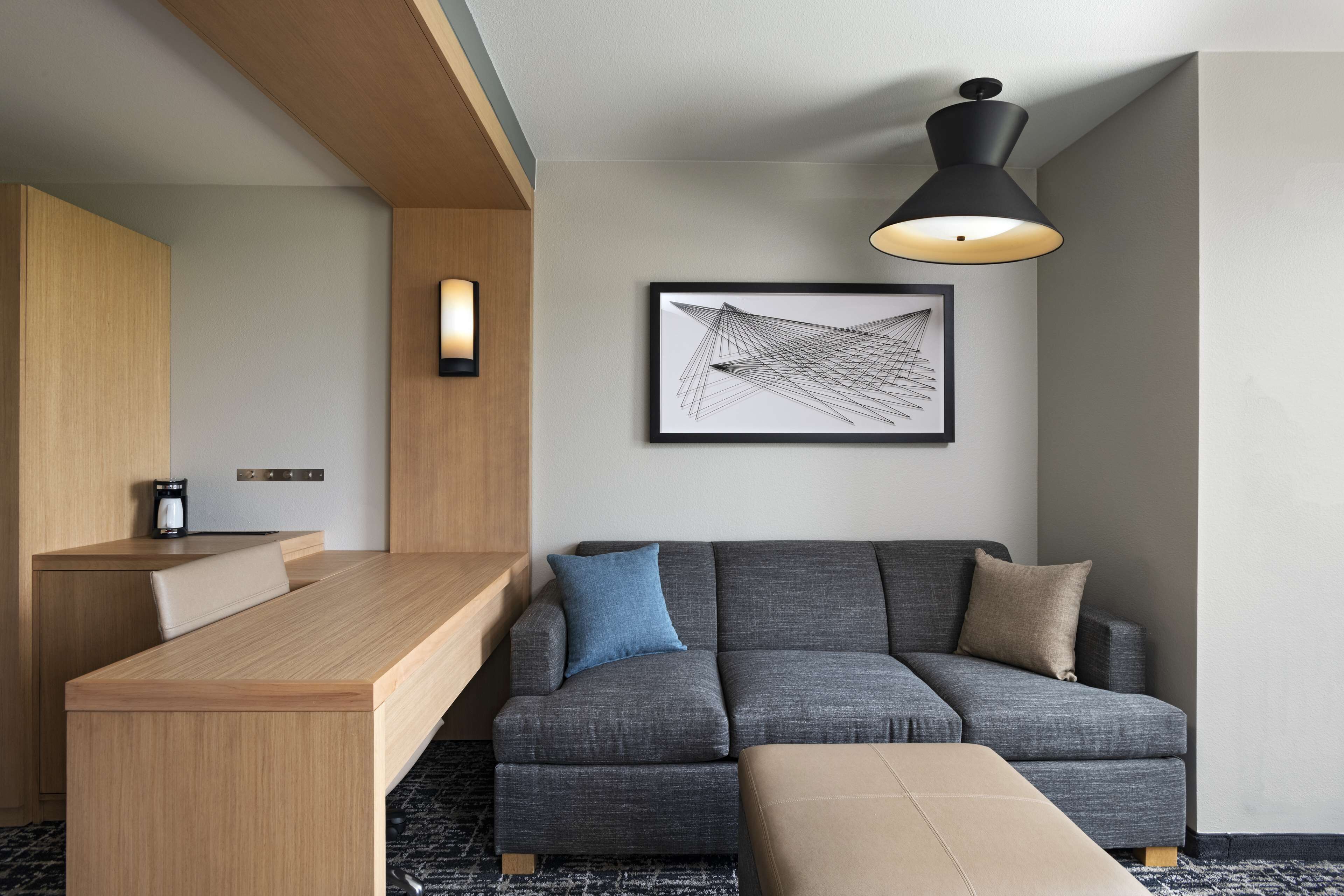 Ein modernes Hotelzimmer mit Schreibtisch aus Holz, einer grauen Couch, einem gerahmten abstrakten Kunstwerk und einer schwarzen Pendelleuchte.