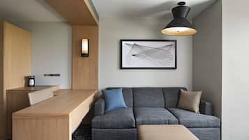 Ein modernes Hotelzimmer mit Schreibtisch aus Holz, einer grauen Couch, einem gerahmten abstrakten Kunstwerk und einer schwarzen Pendelleuchte.