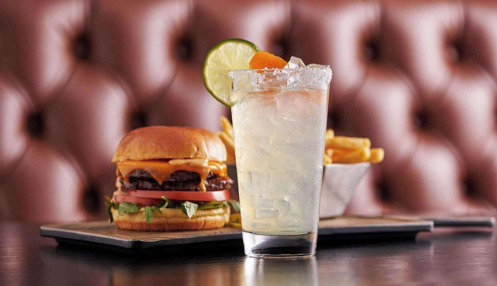 A burger and a glass of iced beverage on a table with a cushioned background.