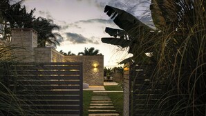 Una entrada con muro de piedra, un camino que lleva a una zona de estar y palmeras.