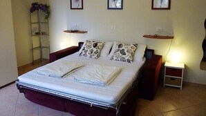 Un lit bien fait, avec un matelas blanc et deux oreillers, une table de chevet avec une lampe, et une étagère fixée au mur sur laquelle est posé un cadre photo.