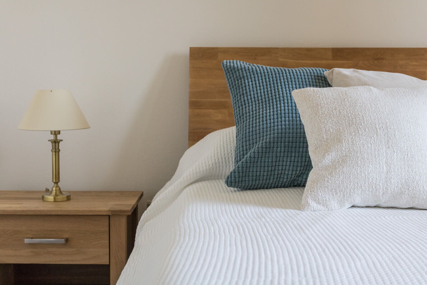 A neatly made bed with white bedding and two pillows, one blue and one white, against a wooden headboard. A brass table lamp is on a wooden nightstand beside the bed.