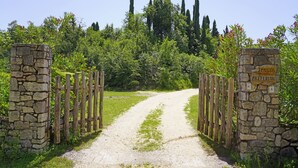 Un chemin de gravier mène à travers une porte en pierre, flanquée de piliers en bois, et s’enfonce dans un écrin de verdure luxuriante, bordé de grands arbres.