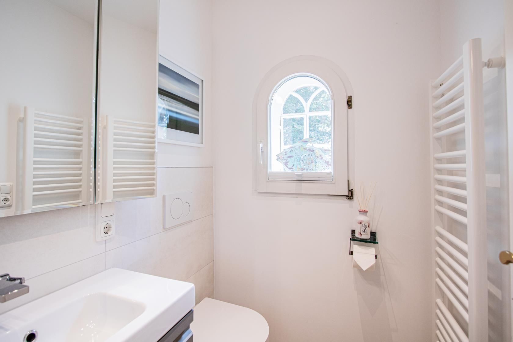 Ein Badezimmer mit einem weißen Waschbecken, einer Toilette und einem Fenster mit Blick auf Bäume.