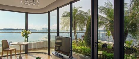A modern living room with a large TV, a dining table, and a view of a body of water and palm trees.